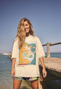 Leende kvinna med långt hår står på en träpir vid havet, iklädd en grafisk oversize skjorta och färgglada rutiga shorts under klar himmel.