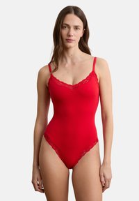 Frau trägt einen eng anliegenden roten Body mit Spitzenbesatz und dünnen Trägern, steht vor einem schlichten weißen Hintergrund.