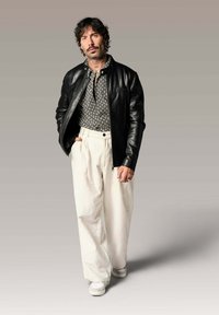 Veste en cuir noire sur une chemise à motifs, pantalon large crème et baskets blanches. Tatouages visibles sur les bras et le cou.