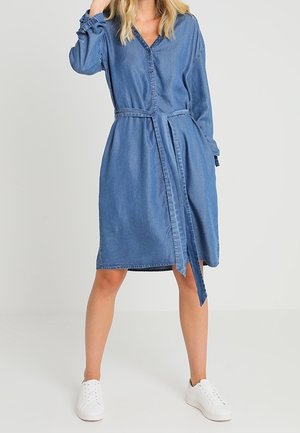 Femme portant une robe-chemise en jean bleu longueur genou avec manches longues, une ceinture à la taille et des baskets blanches, debout sur un fond uni.