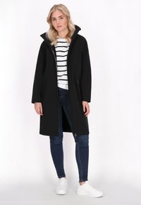 Juodas ilgas paltas su aukštu apykakle, užtrauktuku ir šoninėmis kišenėmis, dėvimas ant dryžuoto marškinėlio, derinamas su tamsiomis džinsomis ir baltais sportbačiais.