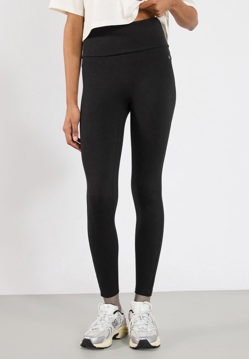 Leggings negre cu talie înaltă, fabricate dintr-un material elastic, având o textură netedă și un design simplu, asortate cu adidași albi și gri.