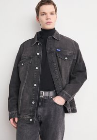 Black denim jacket with two chest pockets, silver buttons, and light fade detail. Worn over a black turtleneck, paired with matching jeans.