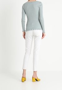 Top a maniche lunghe di un leggero blu ribattuto con bordi scallop, abbinato a jeans bianchi aderenti e mule gialle con tacco a blocco. Vista posteriore.