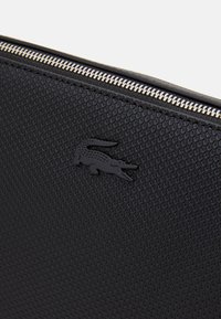 Superficie nera testurizzata con chiusura a zip. Presenta un logo di coccodrillo in rilievo nero, accentuato da un dettaglio della zip color argento.