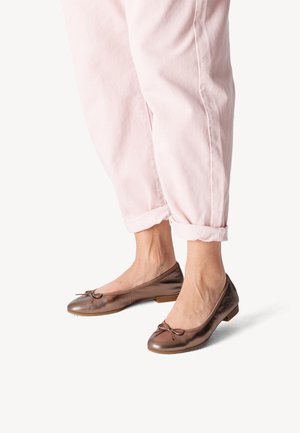 Ben iført lyserøde bukser, der er rullet op, og skinnende bronze ballet flats med små sløjfer mod en hvid baggrund.