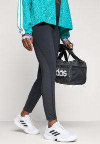 Osoba u crnim tajicama i bijelim Adidas tenisicama, drži crnu Adidas sportsku torbu i nosi jaknu s uzorkom u tirkiznoj boji.