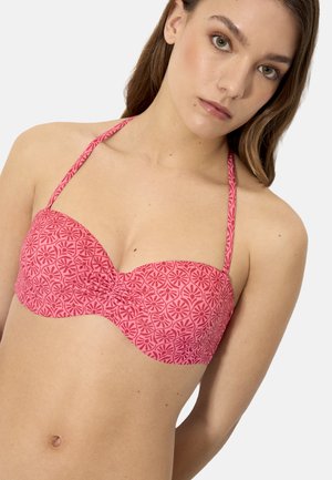 Joven mujer con un top de bikini rosa de tipo halter con un patrón geométrico floral, mirando ligeramente hacia adelante sobre un fondo blanco.