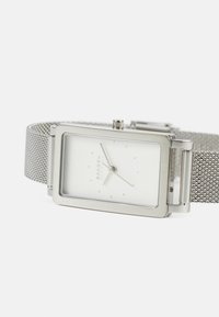 Reloj de pulsera rectangular plateado con esfera blanca, marcadores de hora mínimos y una correa de malla plateada, colocado plano sobre una superficie blanca.