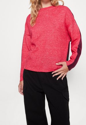 Maglione rosso lavorato a maglia con maniche ampie, caratterizzato da dettagli in rosso scuro sui lati. Il tessuto ha un aspetto leggermente testurizzato. Indossato con pantaloni neri.