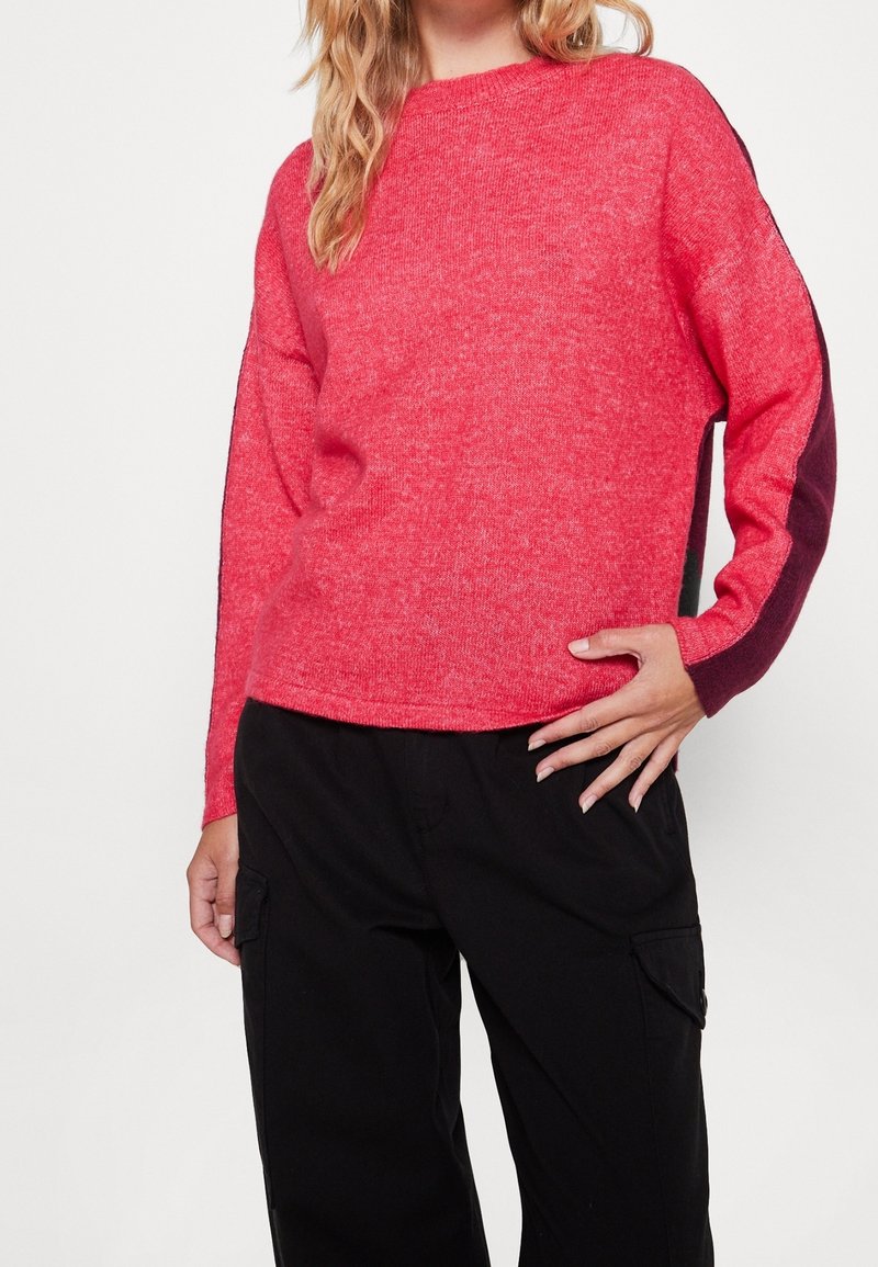 Rot gestrickter Pullover mit weiten Ärmeln, der an den Seiten dunkelrote Akzente aufweist. Der Stoff hat ein leicht strukturiertes Aussehen. Kombiniert mit schwarzen Hosen.