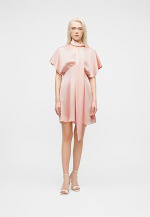 Vrouw draagt een glanzende kort roze jurk met wijde mouwen en een lange sjaaldetail, gecombineerd met witte enkelsandalen met riempje, staand tegen een witte achtergrond.