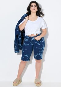 Shorts di jeans con un motivo tie-dye blu, abbinati a una maglietta bianca annodata e a un giubbotto di jeans blu, completati da sandali con plateau beige.