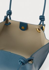 Kék bőrből készült válltáska csomózott fogantyúkkal, bézs belsővel és aranyszínű gombos záródással, felülről nyitva bemutatva.