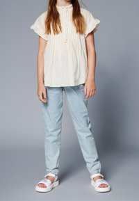 Haut jaune clair à manches courtes avec volants et boutons ; pantalon rayé bleu et blanc ; sandales plates blanches avec des accents imprimés.