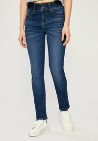 Džínové jeansy v tmavě modré barvě s přiléhavým střihem, vybavené dvojitým zapínáním, pěti kapsami a prošitými detaily. V kombinaci s bílými teniskami.