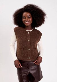 Femme souriante avec de volumineux cheveux bouclés portant un gilet en tricot marron à boutons sur un haut à manches longues crème et un short en cuir marron foncé.