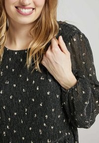 Top en tissu noir avec un motif texturé, featuring des accents floraux dorés et des manches bouffantes. Le tissu a un léger brillant et se resserre au niveau des poignets.