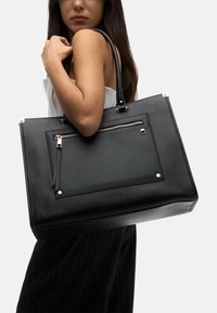 Black tote bag made of textured material, featuring a front zip pocket, gold-tone hardware accents, and dual shoulder straps.