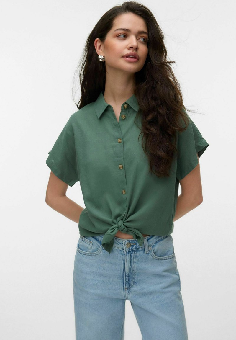 Femme aux longs cheveux foncés portant une chemise verte à manches courtes boutonnée et nouée à la taille ainsi qu'un jean taille haute bleu clair, regardant sur le côté.