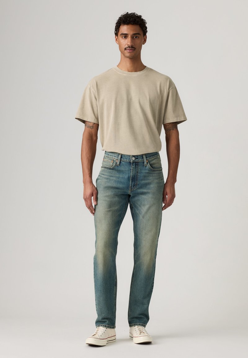 Mand med krøllet hår og overskæg iført beige t-shirt, falmede blå jeans og hvide sneakers står mod en ensfarvet baggrund.