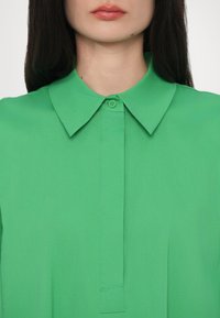 Zielona koszula z kołnierzykiem i zapięciem na guziki, o gładkiej fakturze i dopasowanym kroju, koncentrująca się na prostocie i czystych liniach.
