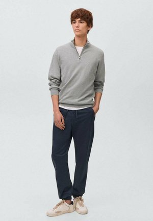Szary sweter zapinany na zamek z prążkowanymi mankietami na białej koszulce, w zestawieniu z granatowymi spodniami i jasnymi sneakersami. Prosty design i casualowy krój.