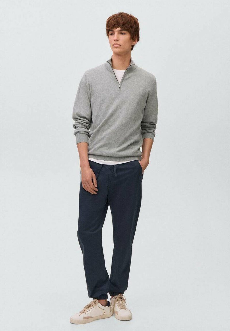 Grå zip-tröja med ribbade muddar över en vit t-shirt, kombinerad med marinblå byxor och ljusa sneakers. Enkelt design och avslappnad passform.