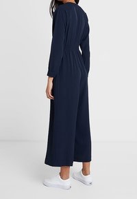 Mujer vestida con un mono azul marino de manga larga con cintura elástica y pantalones anchos, combinado con zapatillas blancas, de pie frente a un fondo liso.