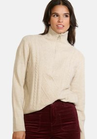 Beeži kabeli kudumisega turtleneck-sviiter, millel on pool tõmblukk, millel on ribatud tekstuur ja teemantmuster, koos burgundi värvi corduroy pükstega.