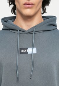 Felpa grigia realizzata in tessuto morbido, dotata di cappuccio regolabile con coulisse, con un'etichetta logo di colore nero e azzurro chiaro sulla parte frontale che recita "JACK & JONES".