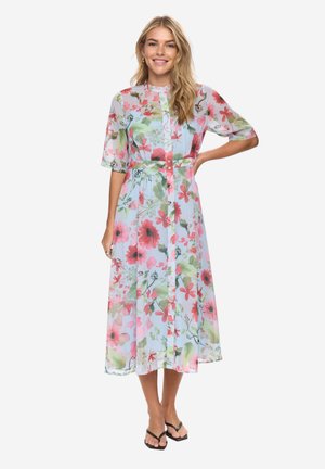 Kvinde smilende, iført en lyseblå midi-kjole med pink og grøn blomstermønster, korte ærmer, knaplukning foran og sorte sandaler med hæl.