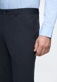 Marineblaue Hose mit glatter Textur und schmaler Passform, ausgestattet mit einem knöpfbaren Bund und seitlichen Taschen, kombiniert mit einem hellblauen Hemd.