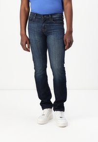 Jeans de mezclilla azul oscuro con corte ajustado, diseño de cinco bolsillos y un ligero desgaste. Combinados con zapatillas blancas y una camisa azul.