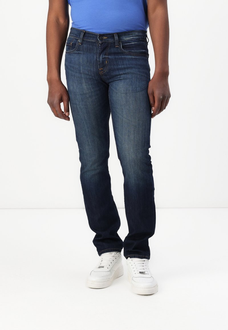 Jeans de mezclilla azul oscuro con corte ajustado, diseño de cinco bolsillos y un ligero desgaste. Combinados con zapatillas blancas y una camisa azul.