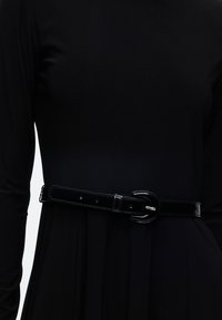 Ceinture en cuir verni noir avec boucle argentée portée sur une robe noire à manches longues à la taille.