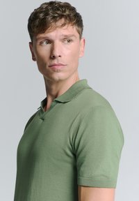 Groen polo-shirt met korte mouwen, ribbelkraag en een soepele textuur; weergegeven op een model dat links kijkt tegen een neutrale achtergrond.