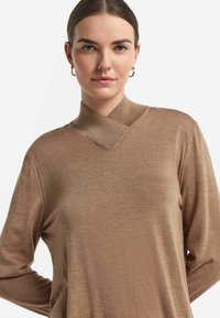 Långärmad brun tröja med hög krage och asymmetrisk halslinje. Tillverkad av mjukt tyg, har den en slät yta och avslappnad passform.