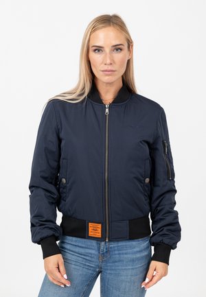 Woman with long blonde hair wearing a navy bomber jacket with black cuffs and light blue jeans, standing against a white background.