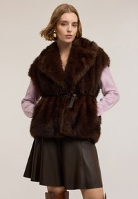 Kobieta w ciemnobrązowej futrzanej kamizelce na jasnofioletowym swetrze, ciemnobrązowej skórzanej spódnicy i brązowych kozaczkach za kolano, stojąca z rękami w kieszeniach.