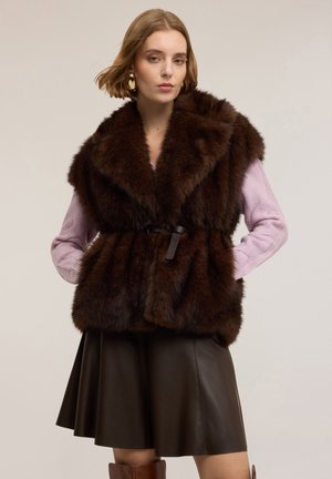 Kobieta w ciemnobrązowej futrzanej kamizelce na jasnofioletowym swetrze, ciemnobrązowej skórzanej spódnicy i brązowych kozaczkach za kolano, stojąca z rękami w kieszeniach.