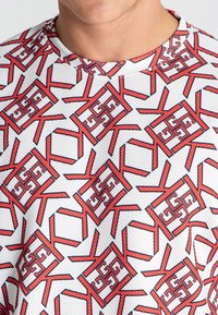 Bijela atletska majica s geometrijskim crvenim uzorkom. Dizajn uključuje međusobno povezane oblike s teksturiranom površinom i okruglim izrezom.