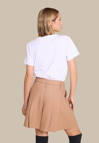 Beige plissert skjørt med tettsittende midje og sidelommer, kombinert med en hvit t-skjorte. Bakgrunnen er i en lys nøytral farge.