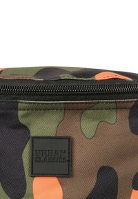 Maskovací taška v odstínech zelené, hnědé a oranžové; obsahuje černý zip a čtvercovou černou nálepku s raženým textem "URBAN CLASSICS."