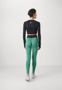 Crop top nero a maniche lunghe con logo; leggings verdi dal design aderente; modello in piedi su sfondo bianco che indossa scarpe da ginnastica bianche.