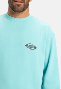 Light blue long-sleeve sweatshirt with a round neck and a small multicolored logo featuring gray and pink accents on the chest.