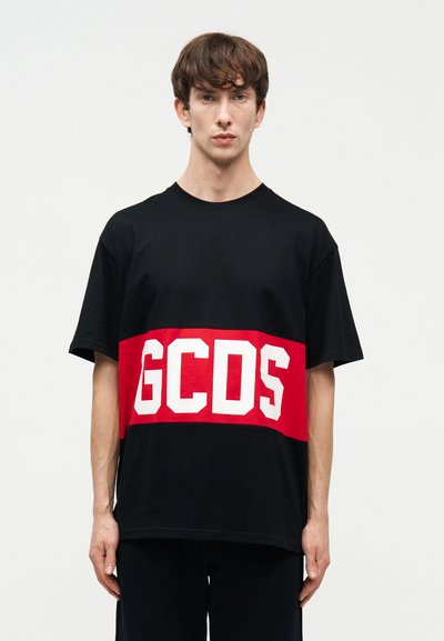 Young man standing facing forward wearing a black T-shirt with a wide red horizontal stripe and white "GCDS" letters across the chest.