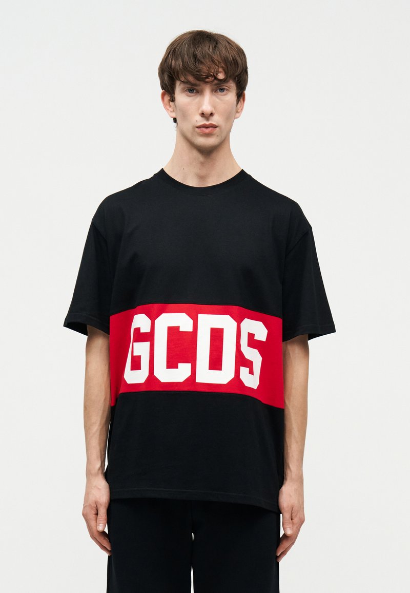 Ung mand, der står med front mod kameraet, iført en sort T-shirt med en bred rød vandret stribe og hvide "GCDS" bogstaver på brystet.