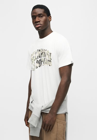 Νέος άνδρας που φοράει λευκό γραφικό T-shirt με το κείμενο "Billionaire Boys Club", ανοιχτό γκρι φούτερ δεμένο γύρω από τη μέση και μπεζ παντελόνι.