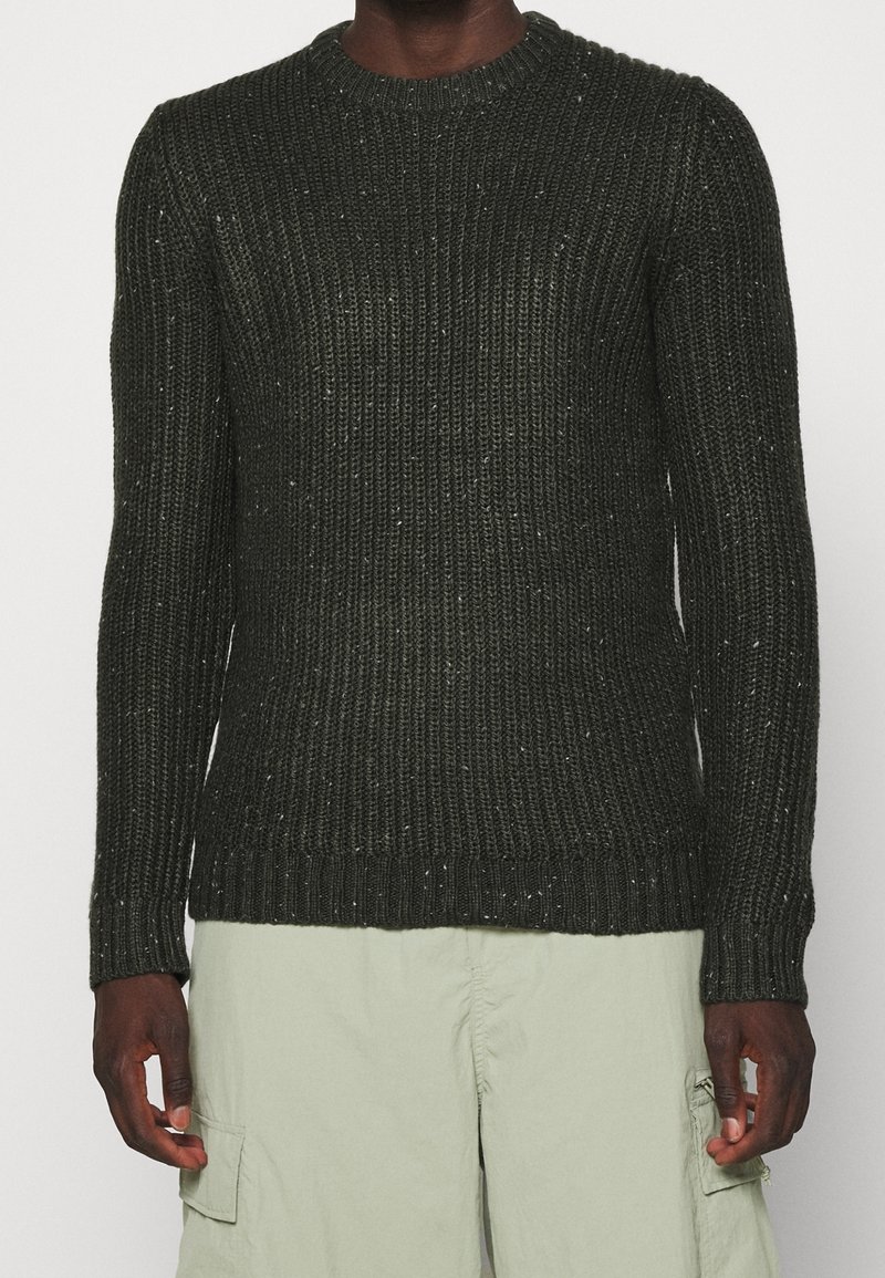 Dunkelgrüner, gerippter Strickpullover, getragen von einer Person mit dunkler Haut, kombiniert mit hellbeigen Cargohosen vor einem schlichten Hintergrund.